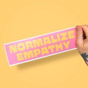 Puede incluir: Una pegatina rectangular rosa con las palabras "NORMALIZE EMPATHY" en letras amarillas. La pegatina está sujeta por una mano con un tatuaje, sobre un fondo amarillo.