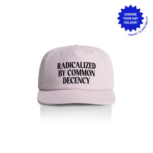 May include: A light pink baseball cap with the text "RADICALIZED BY COMMON DECENCY" in black. The cap has a curved brim and a rounded crown. A blue starburst graphic with the text "CHOOSE YOUR HAT COLOUR!" is in the upper right corner.