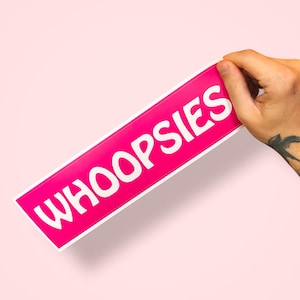 May include: A rectangular sign with the word "WHOOPSIES" in white, bold letters on a bright pink background. The sign has a white border and is held by a hand with a tattoo on the wrist. The background is a soft pink.
