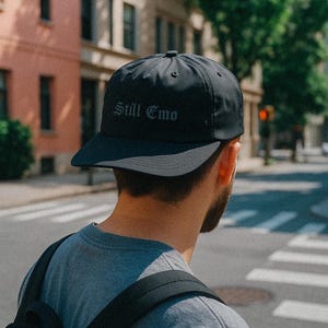 Może przedstawiać: Czarna czapka bejsbolowa z napisem "Still Emo" gotycką czcionką. Czapka ma zakrzywiony daszek i strukturalną koronę. Czapkę nosi osoba z brodą i szarą koszulką.