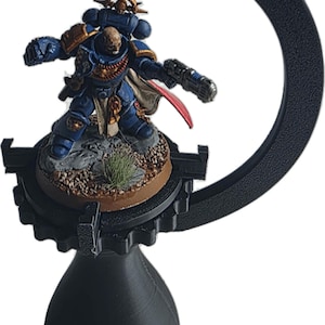 以下が含まれることがあります： 黒色の3Dプリントされたミニチュアフィギュアディスプレイスタンド。スタンドは円形の腕と涙滴型のベースを備えています。青い鎧を着たミニチュアフィギュアが武器を持ってスタンドに展示されています。