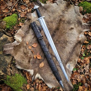 May include: A double-edged steel sword with a black leather-wrapped grip and a matching black leather scabbard featuring a cross-laced design. The sword has a silver-coloured pommel and crossguard, with a central fuller running down the blade.