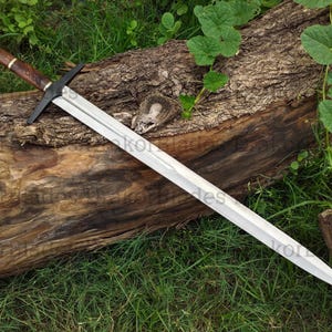 May include: A long, silver sword with a black crossguard and a brown and black handle rests on a weathered log. The sword's blade is sharp and pointed. The background features green grass and foliage.