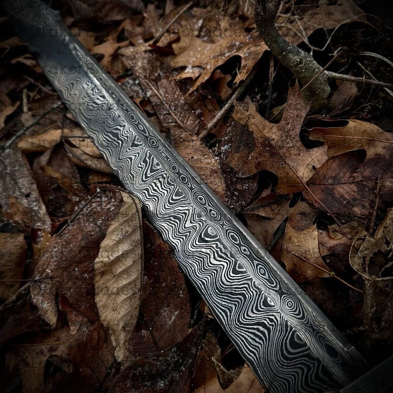 May include: Detailed view of a metal blade with a distinctive patterned surface, likely a sword or knife. The blade features a layered, wavy design in grey and black. It rests amidst a bed of brown autumn leaves and small branches, offering a contrast in textures.