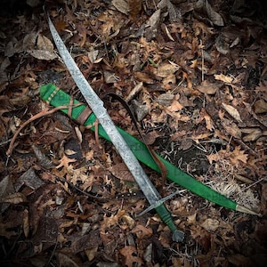 May include: An intricately patterned sword lies on a green scabbard with brown leather straps, set against a bed of autumn leaves. The sword has a crossguard and detailed handle, evoking a medieval or fantasy aesthetic. The blade's length is visible.