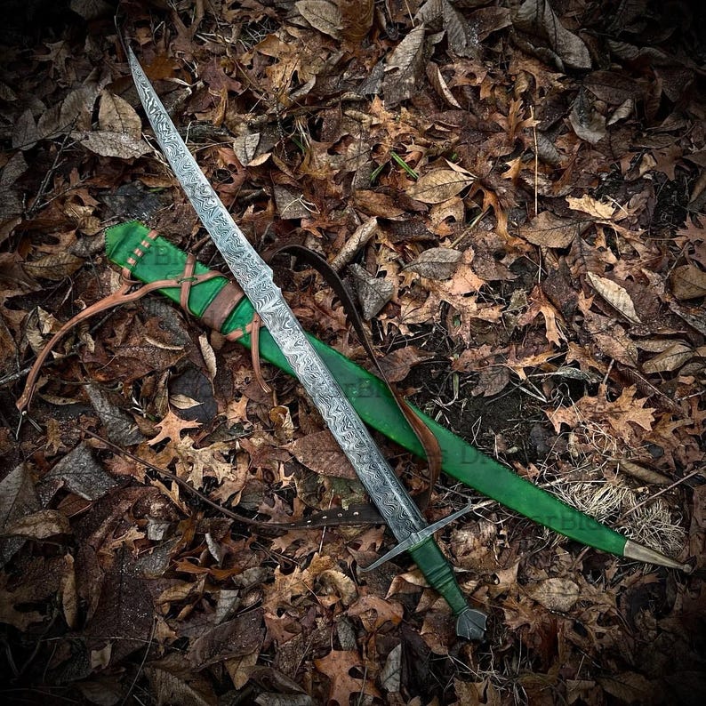 May include: An intricately patterned sword lies on a green scabbard with brown leather straps, set against a bed of autumn leaves. The sword has a crossguard and detailed handle, evoking a medieval or fantasy aesthetic. The blade's length is visible.