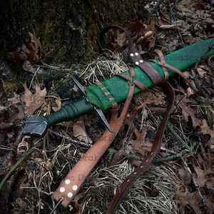May include: A medieval sword with a green leather-wrapped handle and a silver blade. The sword is sheathed in a green leather scabbard with brown leather straps. The scabbard is attached to a brown leather belt. The sword rests on a bed of leaves and moss.