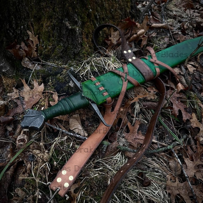 May include: A medieval sword with a green leather-wrapped handle and a silver blade. The sword is sheathed in a green leather scabbard with brown leather straps. The scabbard is attached to a brown leather belt. The sword rests on a bed of leaves and moss.