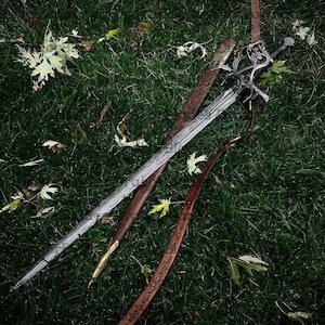 May include: Two swords with ornate silver hilts and patterned blades rest on green grass with fallen leaves. One sword is sheathed in a brown leather scabbard. A second brown leather scabbard and strap are also visible.