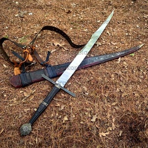 May include: A long, silver sword with a black handle and crossguard, lying on brown pine needles. The sword is sheathed in a leather scabbard with a decorative pattern. A leather strap is also visible.
