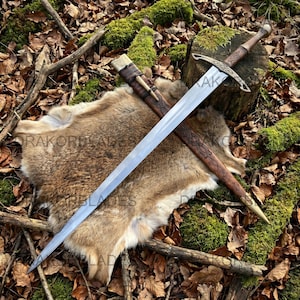 May include: A double-edged steel sword with a brown leather-wrapped hilt and a metallic crossguard featuring intricate knotwork. The sword is accompanied by a matching brown leather scabbard with embossed patterns and a light-colored bone accent.