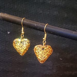 Puede incluir: Un par de pendientes dorados en forma de corazón con textura brillante. Los pendientes cuelgan de ganchos dorados. El fondo es de color oscuro y liso.