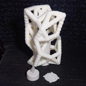 Op de afbeelding: Een witte, abstracte kandelaar met een geometrisch ontwerp. De houder heeft een roosterachtige structuur. Een kleine, witte LED-kaars en het batterijdeksel liggen naast de houder. De kandelaar is gemaakt van een dik, getextureerd materiaal.