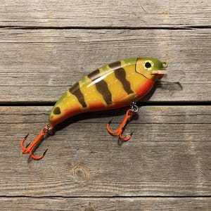 May include: A fishing lure with a yellow and green body featuring dark brown vertical stripes and a bright orange underside. The lure has a clear plastic diving lip at the front, a single eye, and two orange treble hooks attached to the bottom.