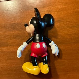 May include: A Mickey Mouse figurine, featuring a black body, ears, and arms, with a red shorts, yellow shoes, and white gloves. The classic cartoon character is posed on a wooden surface.