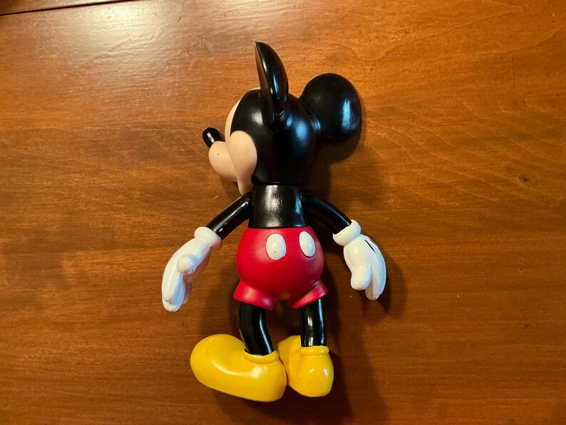 May include: A Mickey Mouse figurine, featuring a black body, ears, and arms, with a red shorts, yellow shoes, and white gloves. The classic cartoon character is posed on a wooden surface.