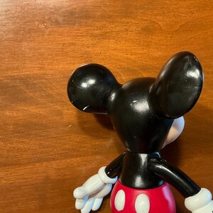 May include: A black Mickey Mouse figurine with white gloves, a red shorts, and a white face. The figurine has large black ears and is positioned on a wooden surface.