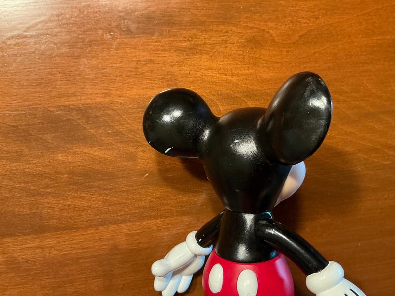 May include: A black Mickey Mouse figurine with white gloves, a red shorts, and a white face. The figurine has large black ears and is positioned on a wooden surface.