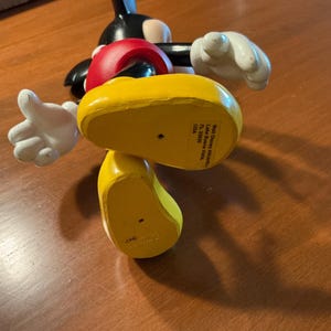 May include: A Mickey Mouse figurine, featuring a black body, red shorts, and yellow shoes. The figure has white gloves and a smiling expression. The bottom of the yellow shoe has text that reads "Walt Disney Attractions, Lake Buena Vista, FL 32830, USA."