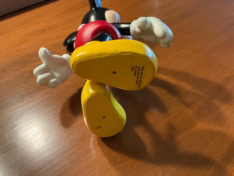 May include: A Mickey Mouse figurine, featuring a black body, red shorts, and yellow shoes. The figure has white gloves and a smiling expression. The bottom of the yellow shoe has text that reads "Walt Disney Attractions, Lake Buena Vista, FL 32830, USA."