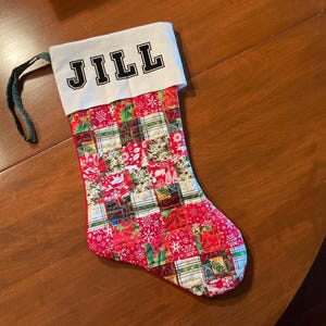 May include: A red and green Christmas stocking with a white cuff that says "JILL" in black letters. The stocking is made of patchwork fabric and has a green ribbon.