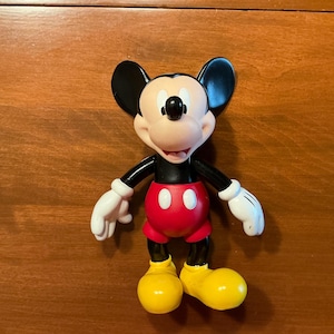 May include: A Mickey Mouse figurine. The figure has a black body, ears, and arms, with white gloves. It wears red shorts with two white buttons and yellow shoes. The face is tan with a black nose and a smiling expression.