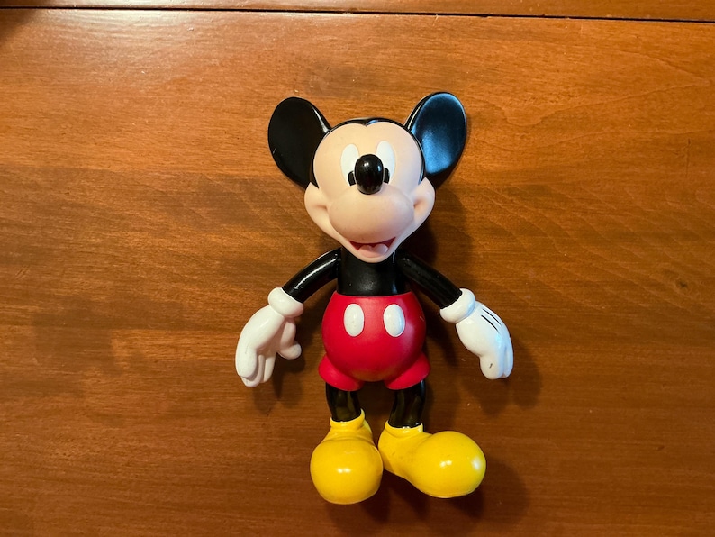 May include: A Mickey Mouse figurine. The figure has a black body, ears, and arms, with white gloves. It wears red shorts with two white buttons and yellow shoes. The face is tan with a black nose and a smiling expression.