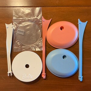 May include: A collection of Barbie doll stands in various colors. The set includes white, pink, and blue stands, along with clear plastic bases and support pieces. The stands are designed to hold dolls upright.