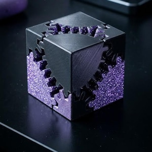 Könnte beinhalten: Ein schwarzer und lila Puzzlewürfel mit Zahnradmuster. Der Würfel hat ein glitzerndes lila Innere und eine matte schwarze Außenseite. Die Zahnräder sind ausgeschnitten, was einen einzigartigen visuellen Effekt erzeugt.