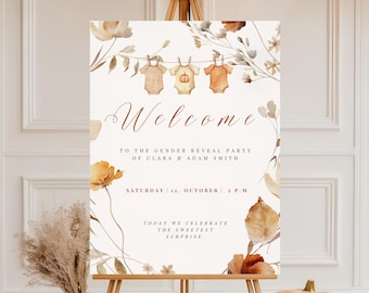 May include: A wooden easel displays a welcome sign for a gender reveal party. The sign features floral illustrations and the text "Welcome" and event details. Pumpkins and fall foliage decorate the scene, creating a warm, autumnal atmosphere.