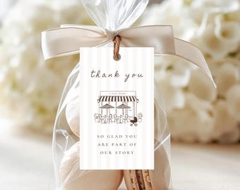 May include: A clear cellophane bag tied with a cream ribbon, containing several macarons. A cream and white striped tag reads "thank you" with a cafe illustration and the text "SO GLAD YOU ARE PART OF OUR STORY". White flowers are in the background.