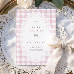 May include: A rectangular baby shower invitation card featuring a light pink and white gingham pattern. The center has a white oval frame with a delicate white lace border, a small bow icon, and text reading: Please join us for a baby shower celebrating mama-to-be Elsie Carter, Sat 10 Oct, at three o'clock in the afternoon, the garden coffee room, Rosewood.