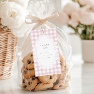 May include: A cellophane bag filled with chocolate chip cookies, tied with a beige ribbon. A pink gingham tag with the words "THANK YOU" and a decorative border is attached. White flowers in a wicker basket are in the background. Cookies are visible.