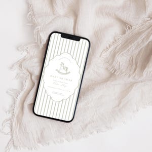 May include: A smartphone screen shows a baby shower invitation with a rocking horse design. The invitation has vertical stripes and a decorative border. The text reads "Baby Shower" and includes event details. The phone is on cream-coloured fabric.