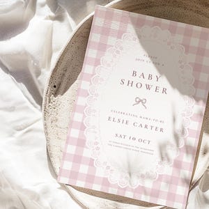 May include: A rectangular baby shower invitation card featuring a light pink and white gingham pattern. The center displays a white oval frame with a delicate lace border, containing the text: Please join us for a baby shower, Elsie Carter, Sat 10 Oct.
