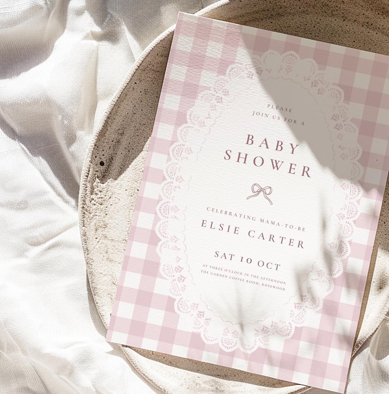 May include: A rectangular baby shower invitation card featuring a light pink and white gingham pattern. The center displays a white oval frame with a delicate lace border, containing the text: Please join us for a baby shower, Elsie Carter, Sat 10 Oct.