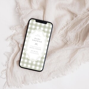 Puede incluir: Un tel&eacute;fono inteligente muestra una invitaci&oacute;n a un baby shower. La pantalla presenta un patr&oacute;n de cuadros vichy verde salvia y blanco con un marco ovalado blanco similar al encaje. El texto de la invitaci&oacute;n dice "Baby Shower" y "Elsie Carter".