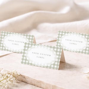 May include: Three folded rectangular place cards featuring a sage green and white gingham pattern. Each card displays a central white oval frame with intricate lace-style detailing and the black printed text YOUR CUSTOM LABEL.