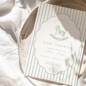 May include: Baby shower invitation with a rocking horse illustration. The invitation has a scalloped border and vertical stripes in a soft green hue. Text includes "Baby Shower" and event details for Elsie Jaye.