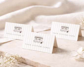 May include: Three folded white place cards with thin vertical light gray stripes. Each card features a dark brown line art illustration of a cafe storefront with tables, umbrellas, and the text "YOUR CUSTOM LABEL" printed in a simple serif font.