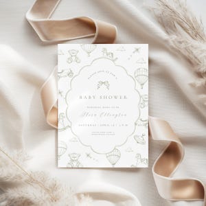 May include: Baby shower invitation with a decorative border and a pattern of toys, hot air balloons, and kites. The invitation text reads "Baby Shower" and includes the honoree's name and event details. A beige ribbon and dried grass are in the background.