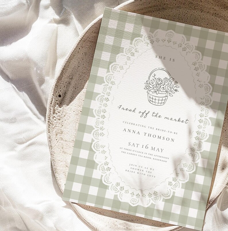 May include: A wedding invitation with a green and white gingham pattern background. The invitation features a floral basket illustration and the text "Fresh off the market". The invitation is on a white plate.