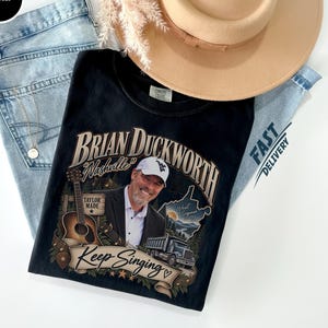 May include: A black t-shirt featuring a graphic print of a man in a suit and cap, an acoustic guitar, a dump truck, and the text Brian Duckworth Nashville Keep Singing. The shirt is displayed with light blue denim jeans and a tan wide-brimmed hat.
