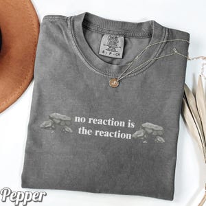 May include: A gray Comfort Colors t-shirt with the phrase "no reaction is the reaction" printed in white. A silver necklace with a pendant is draped across the shirt. The word "Pepper" is in the bottom left corner.