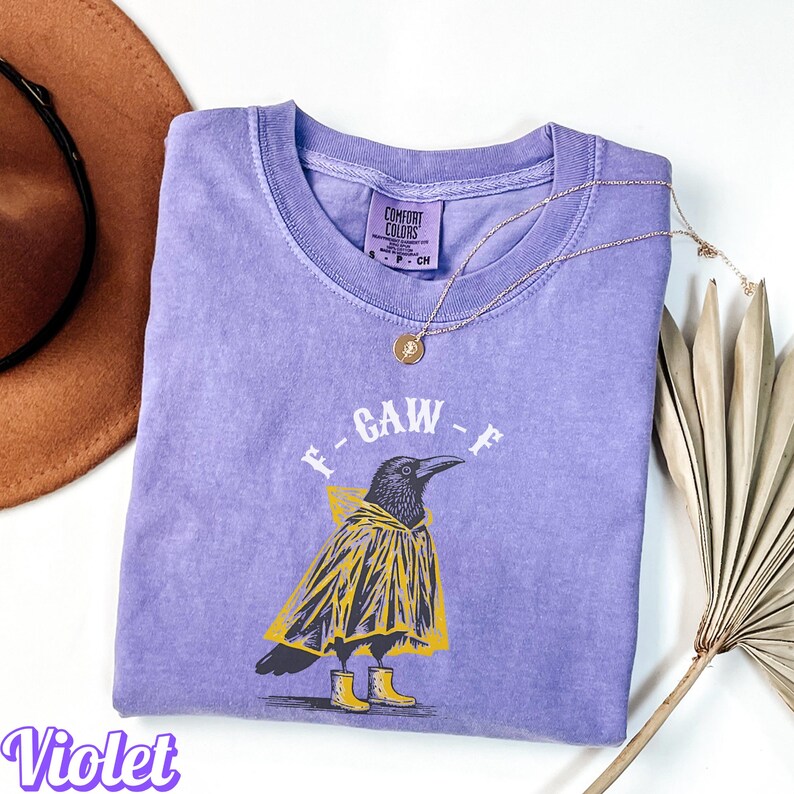 May include: A violet Comfort Colors t-shirt featuring a graphic of a crow wearing a yellow raincoat and boots. The text "F-GAW-F" is above the crow. A gold necklace with a pendant is on the shirt.