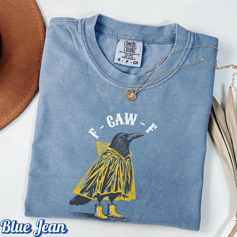 May include: Blue jean colored t-shirt with a graphic of a crow wearing a yellow raincoat and boots. The shirt has the text "F-GAW-F" printed on it. A gold necklace with a pendant is draped across the shirt.