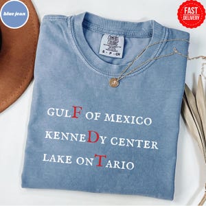 May include: A blue jean colored short-sleeve t-shirt with a crew neckline. The shirt features white text that reads GULF OF MEXICO, KENNEDY CENTER, and LAKE ONTARIO, with the letters F, D, and T highlighted in red. A gold-toned necklace rests on top.