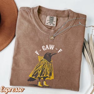 May include: Espresso-colored t-shirt featuring a graphic of a crow wearing a yellow raincoat and boots, with the text "F-GAW-F". A silver necklace with a pendant is draped across the shirt. The shirt has a Comfort Colors tag.