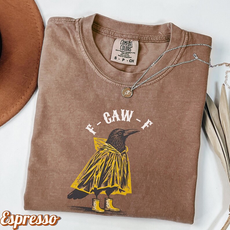 May include: Espresso-colored t-shirt featuring a graphic of a crow wearing a yellow raincoat and boots, with the text "F-GAW-F". A silver necklace with a pendant is draped across the shirt. The shirt has a Comfort Colors tag.