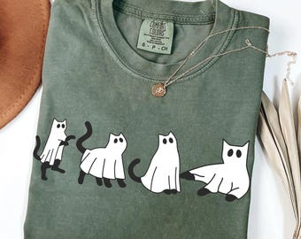 May include: A moss green t-shirt featuring a graphic of four white ghost cats with black accents. The shirt has a crew neck and short sleeves. A gold necklace with a pendant is draped across the shirt.